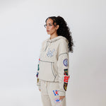 Female model wearing a theW.O.A. Rugby Club Sweat suit in Sandstone with multiple embroidered patches on the sleeves and pant legs on a white background