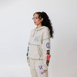 Female model wearing a theW.O.A. Rugby Club Sweat suit in Sandstone with multiple embroidered patches on the sleeves and pant legs on a white background