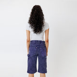 Rear view of a Female model wearing a white t-shirt and Wisdom of age canvas carpenter shorts in purple color on a light gray background