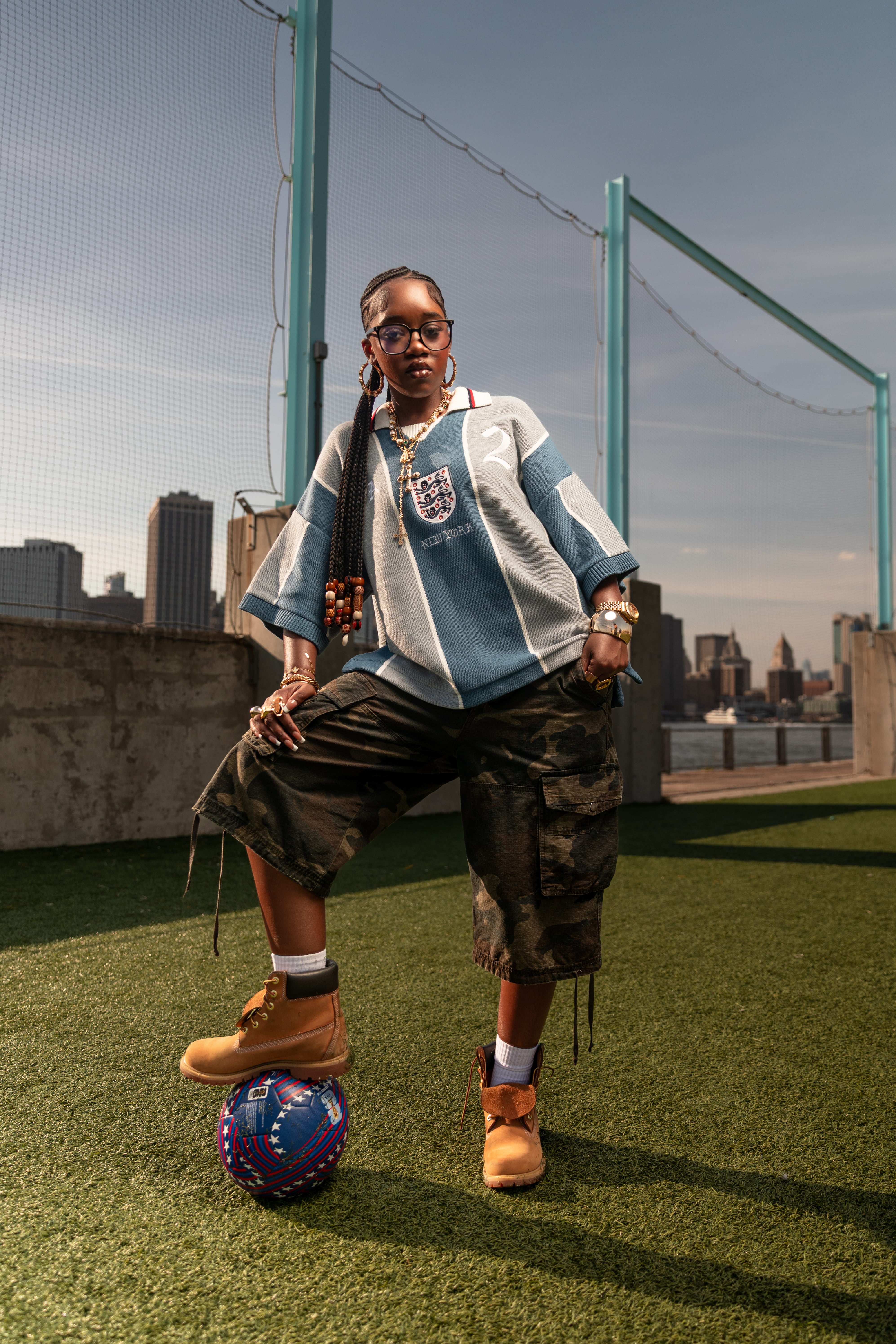African American Female Model standing on a sports field Wearing The Wisdom Of Age Knit Soccer Jersey Grey Stripe with the New York city skyline in the background. She is standing on a World Cup Ball