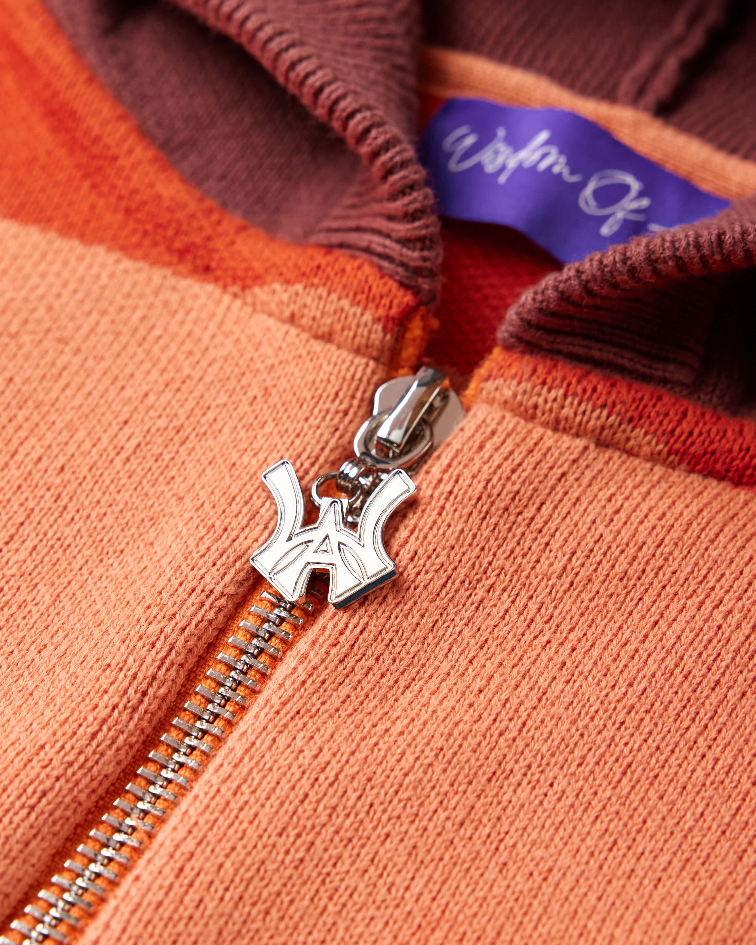 Close Up on the Front view of the WOA knit hoodie. WA logo on the zipper with a chrome finish. Purple tag with Wisdom Of Age written in Signature font.