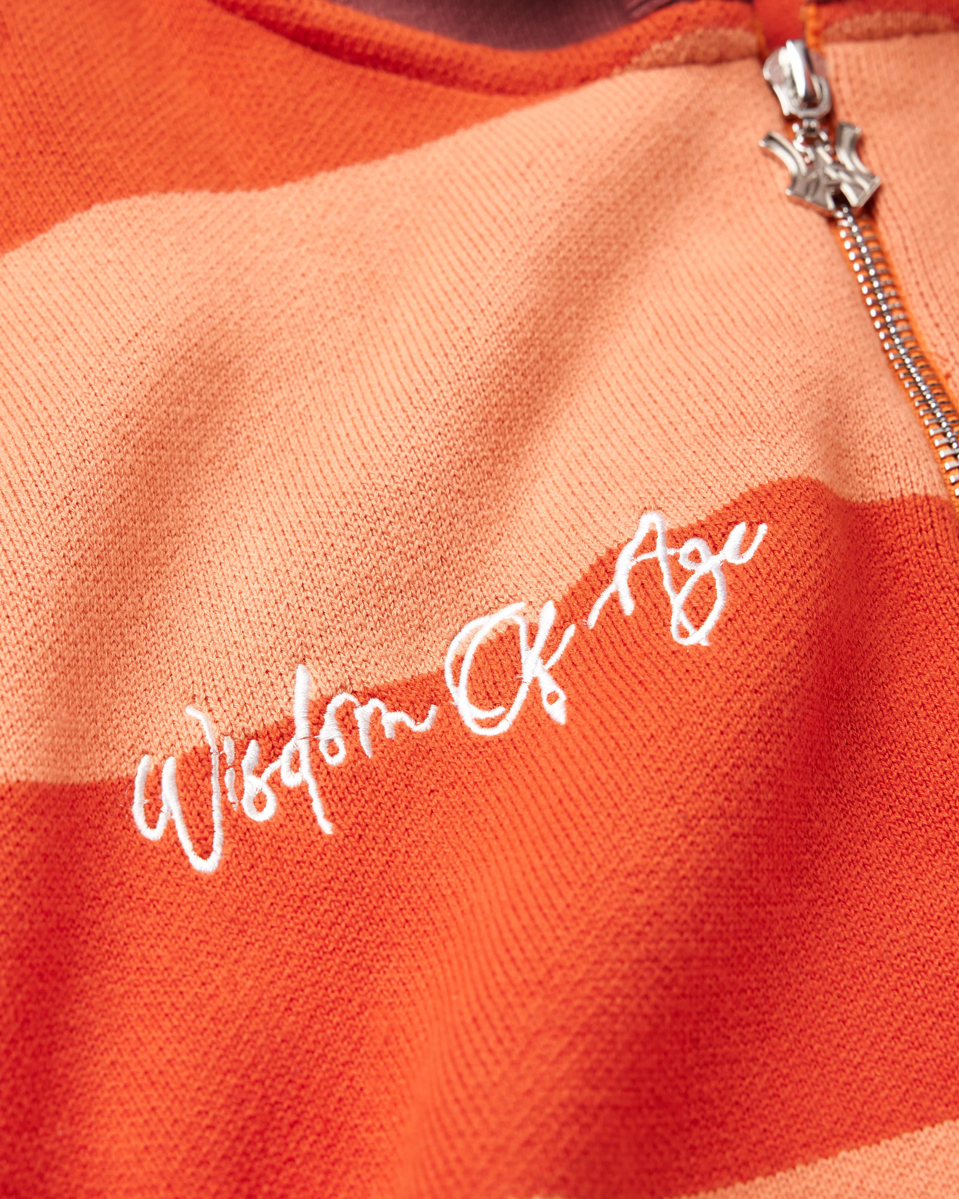 Close Up on the Front view of the WOA knit hoodie. Wisdom Of Age embodied on the right chest in between the alternating orange stripes. Chrome finish WA logo zipper visible on the top right.