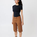 Side view of a Female model wearing a black t-shirt and Wisdom of age canvas carpenter shorts in brick color on a light gray background