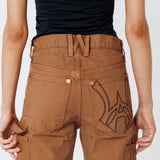 Rear close up view of a Female model wearing a black t-shirt and Wisdom of age canvas carpenter shorts in brick color on a light gray background