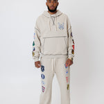 male model wearing a the W.O.A. Rugby Club Sweat suit in Sandstone with multiple embroidered patches on the sleeves and pant legs on a white background
