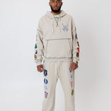 male model wearing a the W.O.A. Rugby Club Sweat suit in Sandstone with multiple embroidered patches on the sleeves and pant legs on a white background