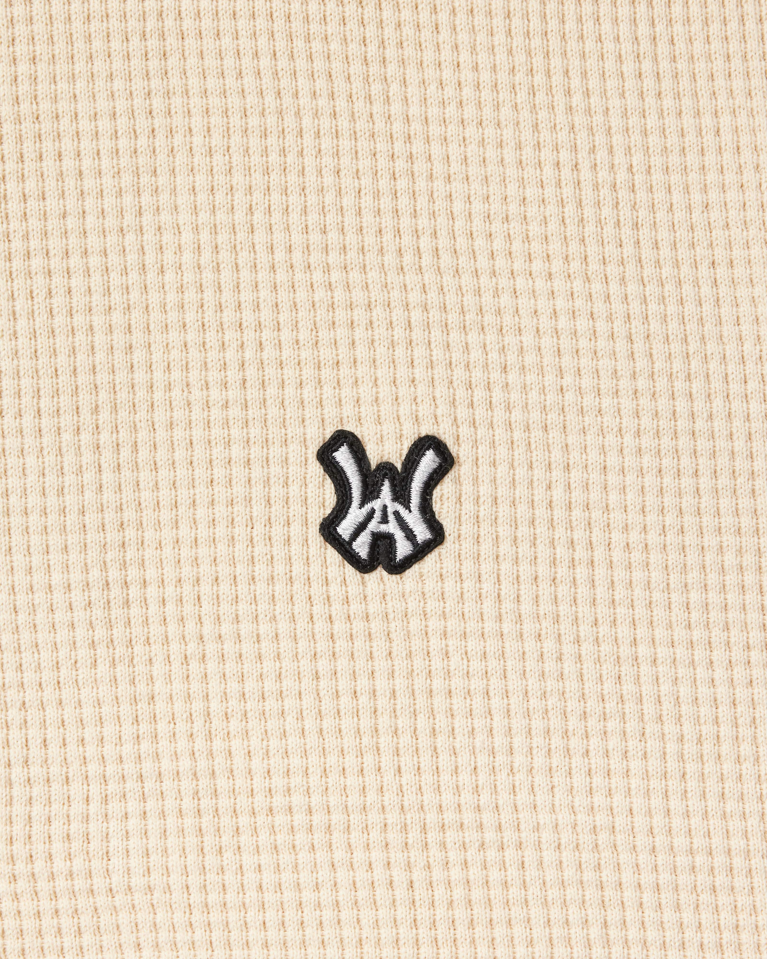 Close up view of the Wisdom Of Age knit thermal shirt in cream. WA Logo embroidered in white with a black outline.