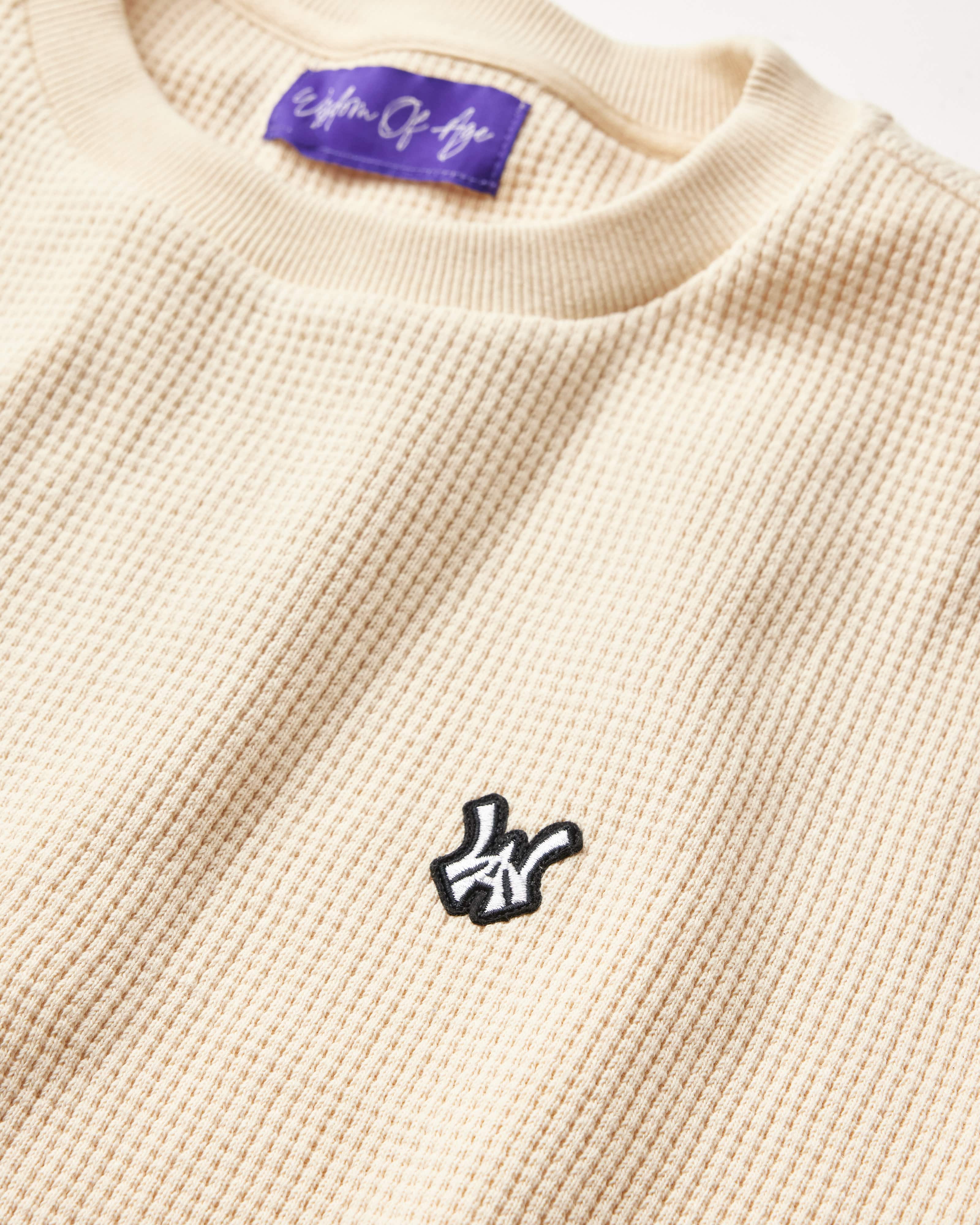 Close Front view of the Wisdom Of Age knit thermal shirt in cream. WA logo embroidered on the left chest in white with a black outline. Purple tag within the collar with Wisdom Of Age written in signature font.