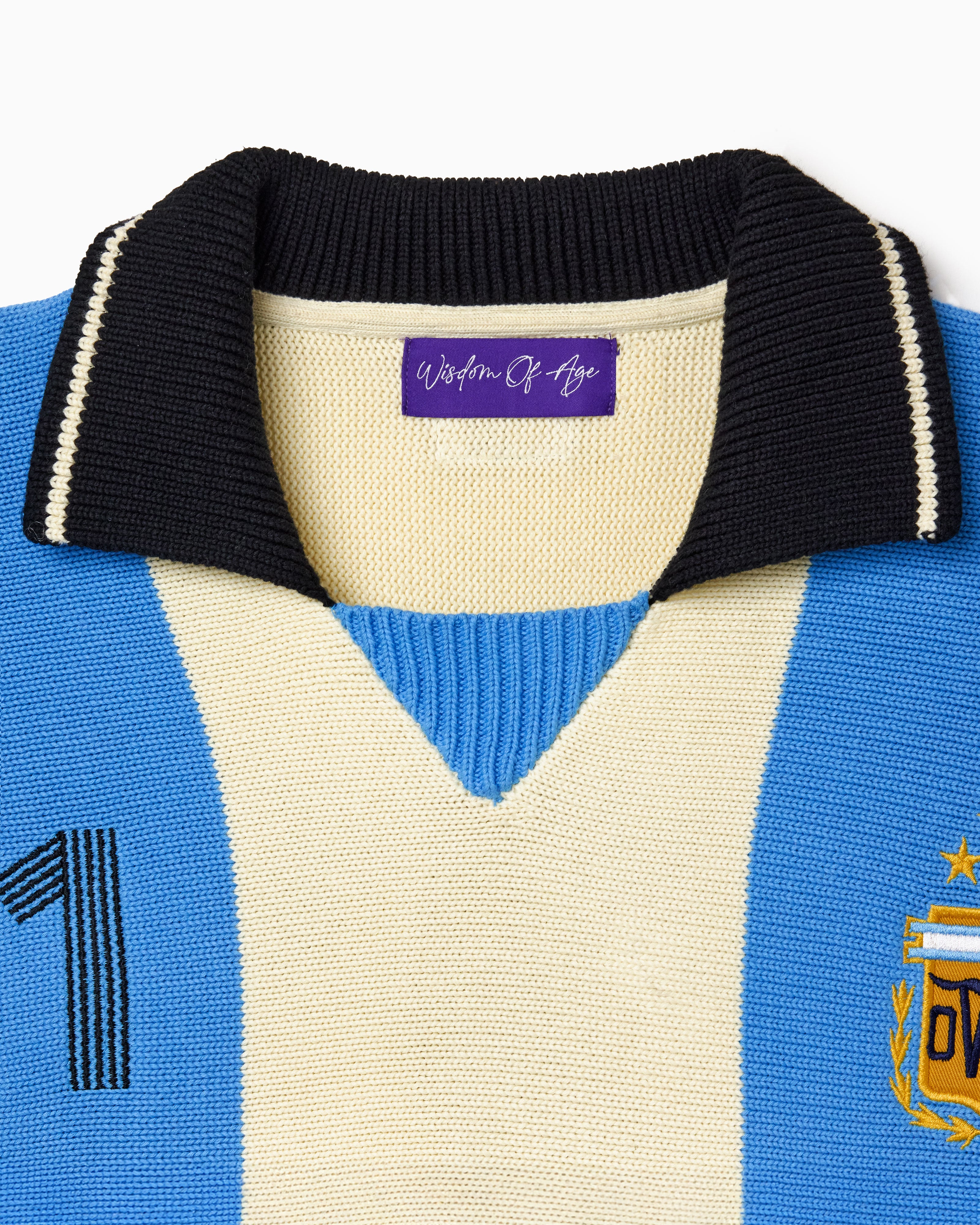 Close-up of a blue and beige knitted soccer jersey with the Wisdom Of Age label on a white background