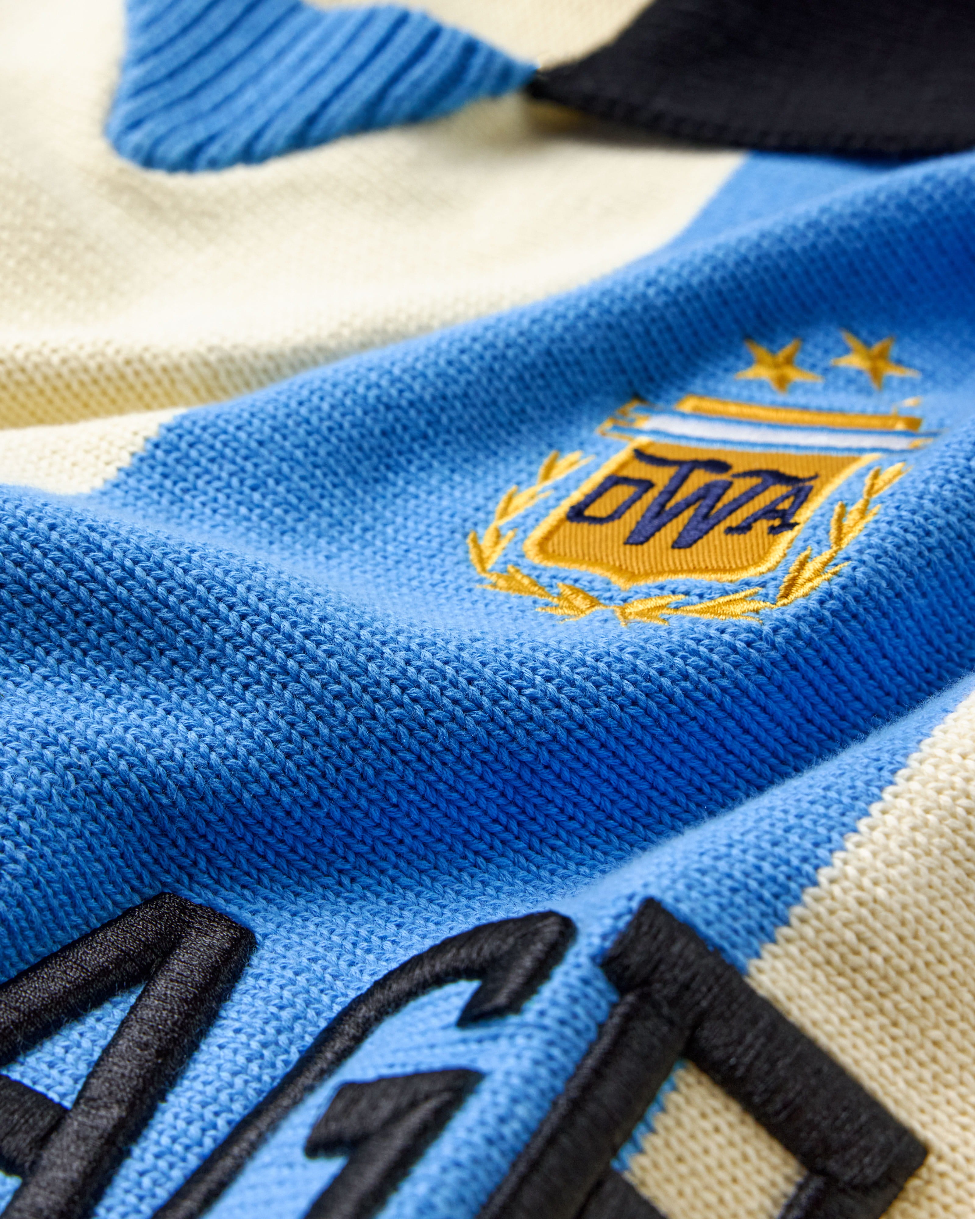 Close-up of The Wisdom Of Age Knit Soccer Jersey Celeste with a logo and text on a blue and beige background