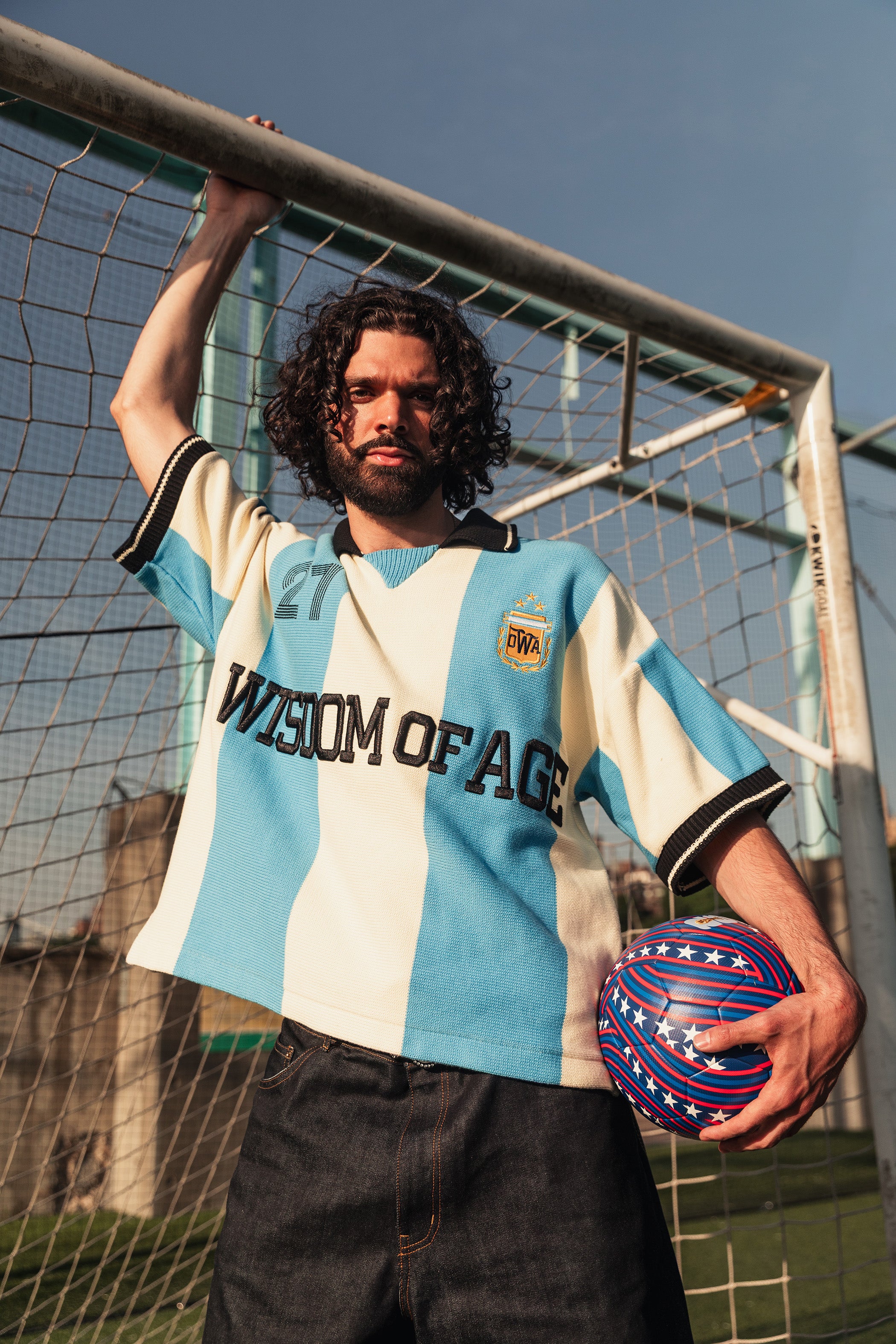 Spanish Model wearing Knit Wisdom Of Age Knit Soccer jersey with 'Wisdom of Age' text, holding a World Cup ball on a soccer field.