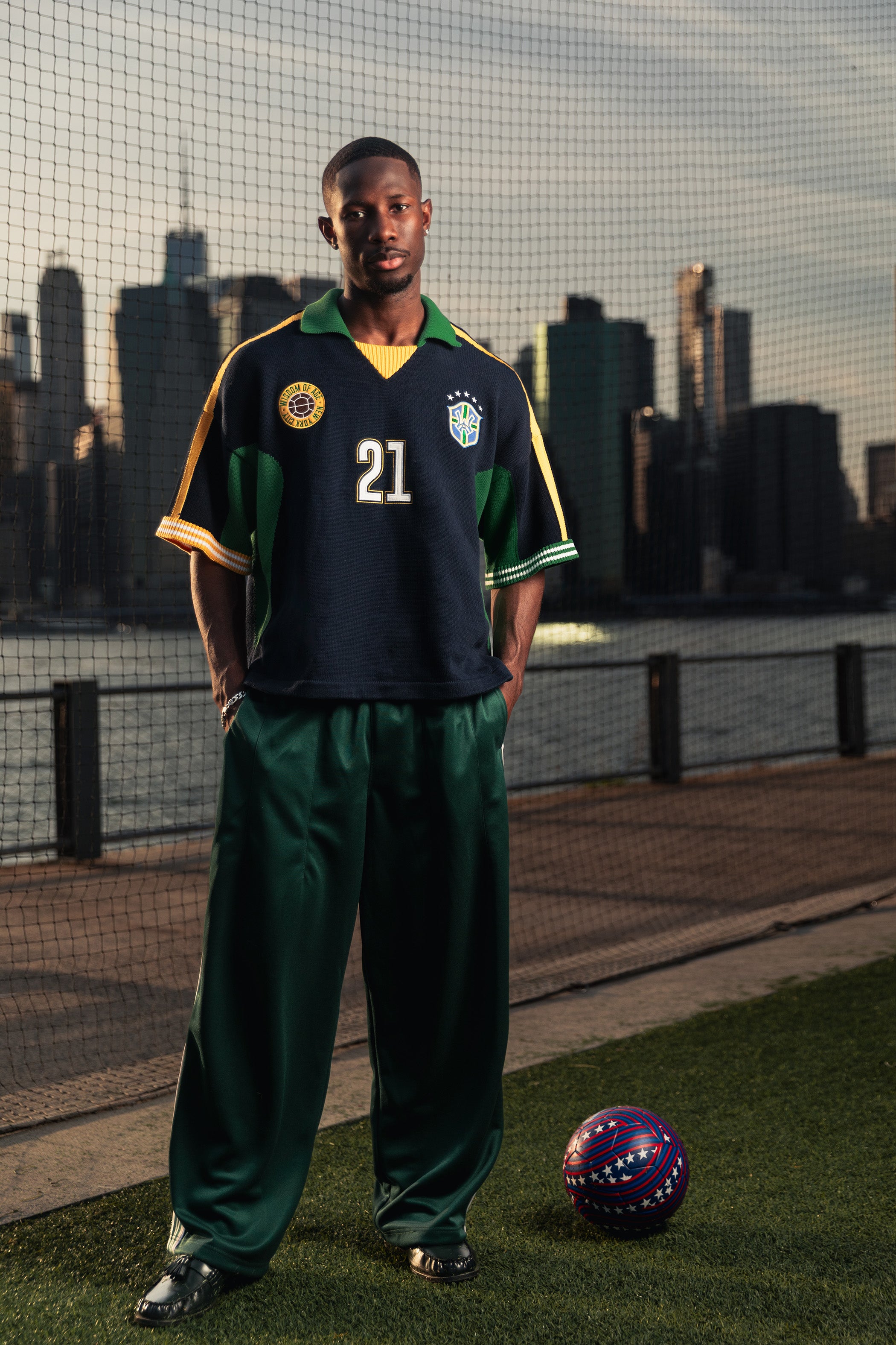 African Model wearing the Knit Wisdom Of Age Soccer jersey Rio with The New York city skyline in the background. He is standing next to a World Cup Ball.